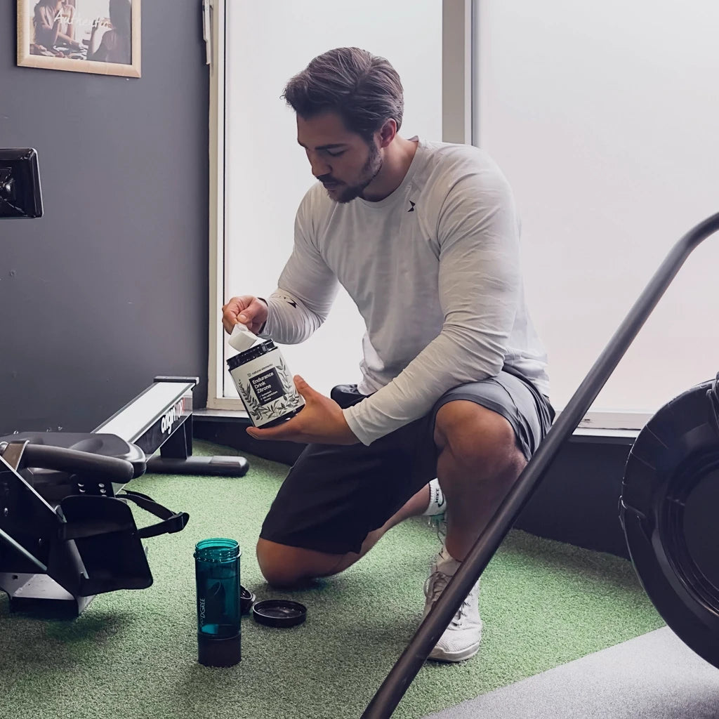 Ein Mann kniet auf dem Boden eines Fitnessstudios und schöpft Pulver aus einem Behälter in eine Shaker-Flasche neben einem Rudergerät.