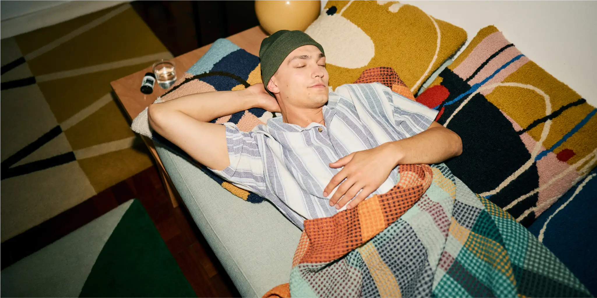 Eine Person mit gestreiftem Hemd und Beanie liegt mit einer bunten Decke zugedeckt auf einem Sofa, hat die Augen geschlossen und den Arm hinter dem Kopf abgelegt.